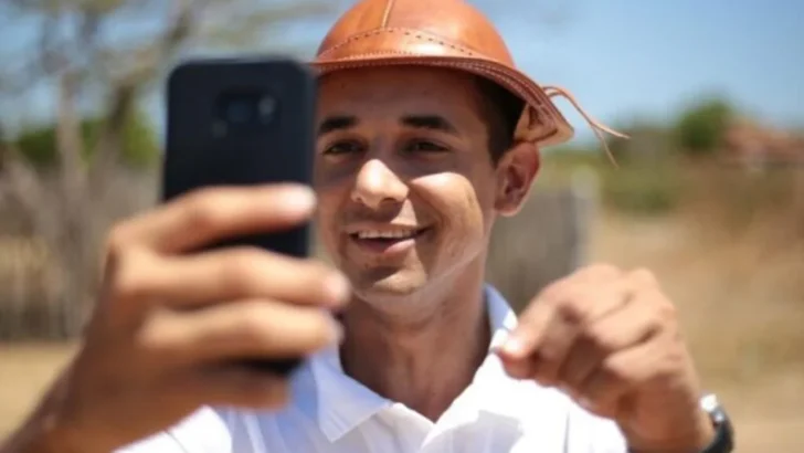 Allyson Bezerra aparece sorrindo, usando camisa polo branca e chapéu de couro, enquanto segura um celular para fazer uma gravação ao ar livre
