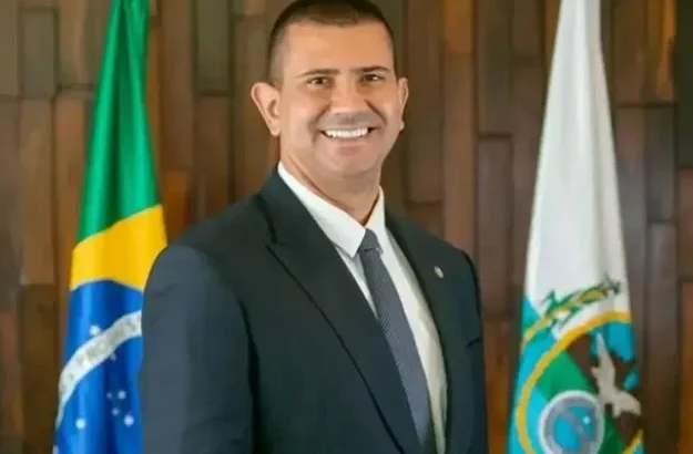 Homem de terno escuro e gravata cinza, sorrindo diante de bandeiras do Brasil e do Estado do Rio de Janeiro