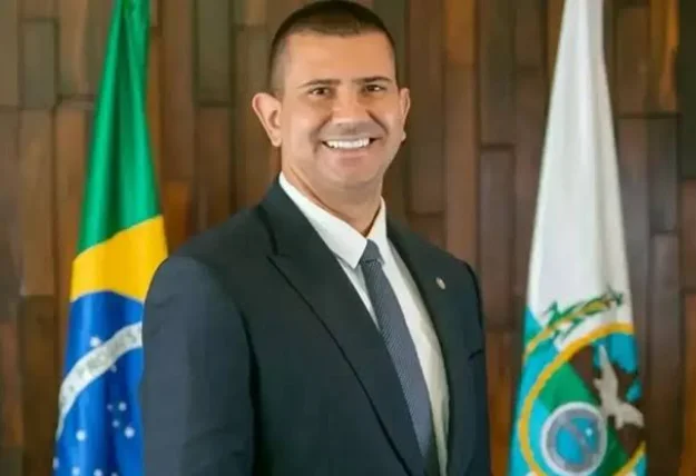 Homem de terno escuro e gravata cinza, sorrindo diante de bandeiras do Brasil e do Estado do Rio de Janeiro