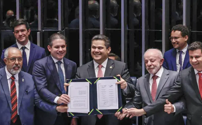 Grupo de autoridades de terno posa para fotografia no Congresso Nacional, enquanto duas pessoas seguram uma pasta aberta com documentos e outras fazem gestos de comemoração