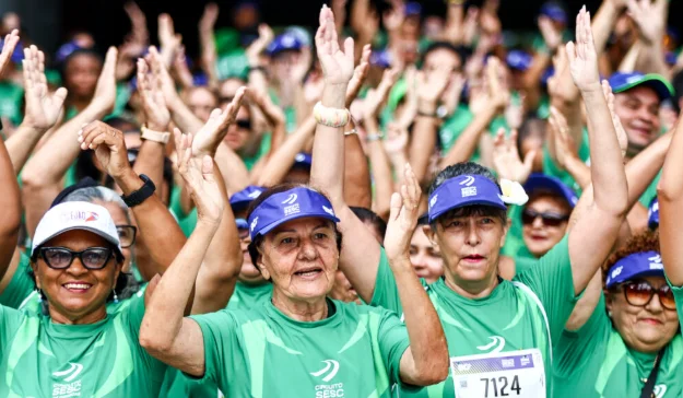 Grupo de participantes de corrida de rua, vestidos com camisetas verdes e bonés azuis, levanta os braços durante evento esportivo do Sesc
