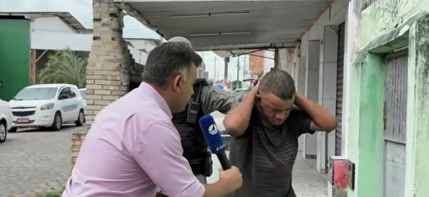 Homem de camiseta escura aparece com as mãos próximas à cabeça enquanto é entrevistado por um repórter que segura um microfone azul; um policial está ao lado