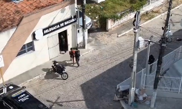 Imagem aérea mostra a fachada da Delegacia de Macaíba, com uma motocicleta estacionada, pessoas na rua e parte de uma viatura da Polícia Civil