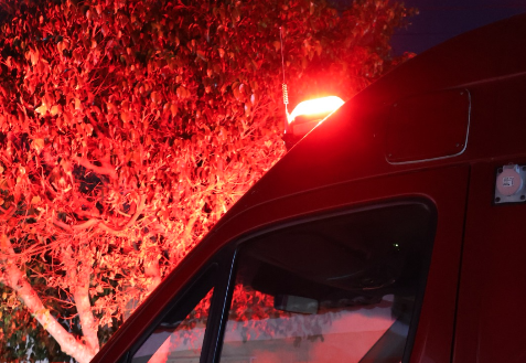 Imagem noturna mostra parte de um veículo de emergência com luz vermelha acionada, iluminando uma árvore próxima