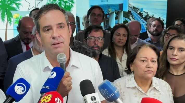 Homem de camisa branca fala ao microfone diante de jornalistas, cercado por várias pessoas. Uma mulher de camisa clara aparece ao seu lado, enquanto microfones de emissoras de comunicação são posicionados à frente dos participantes