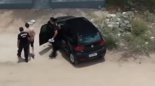 Imagem registrada de cima mostra dois agentes policiais conduzindo um homem próximo a um carro preto estacionado em uma área de terra. Uma das portas do veículo está aberta