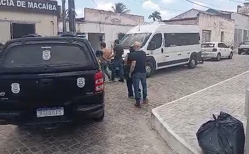 Imagem mostra pessoas próximas a uma viatura preta da Polícia Civil e a uma van branca, estacionadas em uma rua de paralelepípedos, em frente a um prédio com identificação de Macaíba