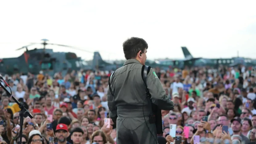 Artista com roupa de estilo militar aparece de costas para uma multidão durante apresentação ao ar livre, com aeronaves militares ao fundo e espectadores registrando o momento com celulares