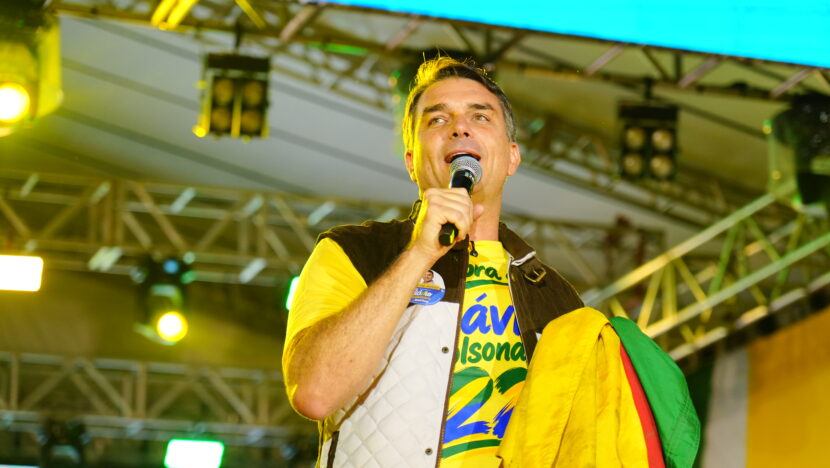 Flávio Bolsonaro fala ao microfone em palco com bandeira brasileira nos ombros