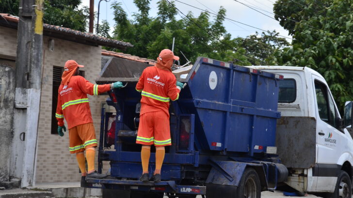 Dois trabalhadores com uniformes laranja na traseira de um caminhão de coleta de lixo em uma rua residencial