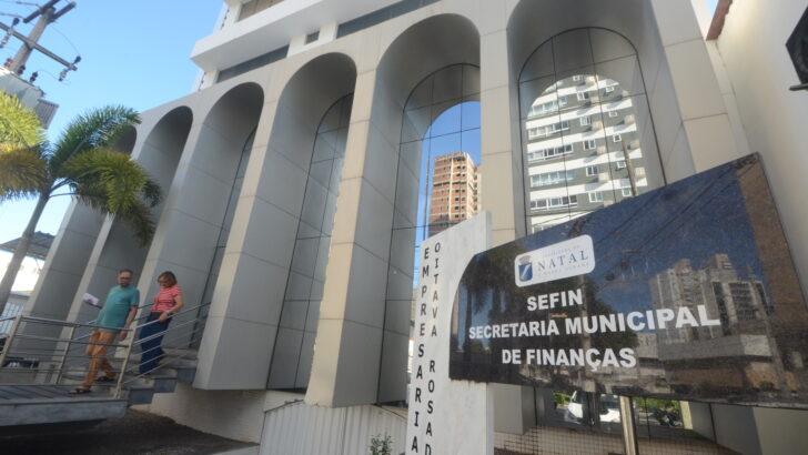 Fachada da Secretaria Municipal de Finanças de Natal, com placa de identificação da Sefin e pessoas nas escadas de acesso ao prédio