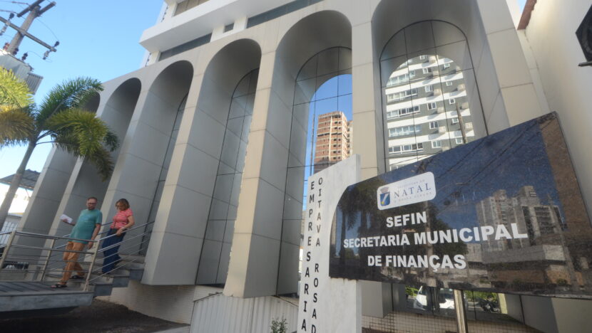 Fachada da Secretaria Municipal de Finanças de Natal, com placa de identificação da Sefin e pessoas nas escadas de acesso ao prédio