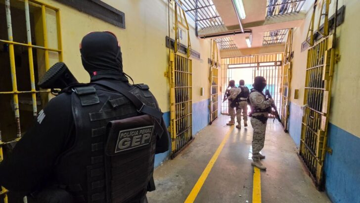 Agentes de segurança com coletes e armamentos em corredor de presídio, com portas de grades amarelas, paredes azuis e identificação da Polícia Penal GEP