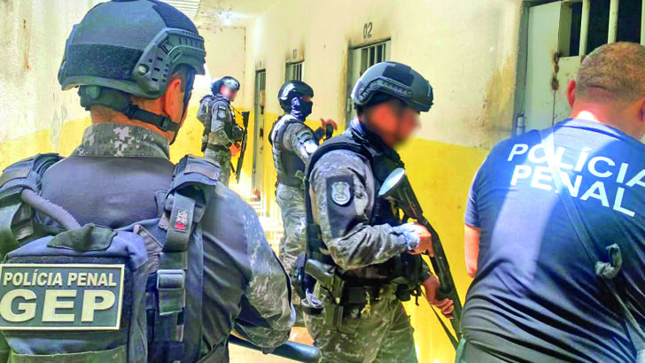 Policiais penais com equipamentos de proteção realizam inspeção em corredor de unidade prisional