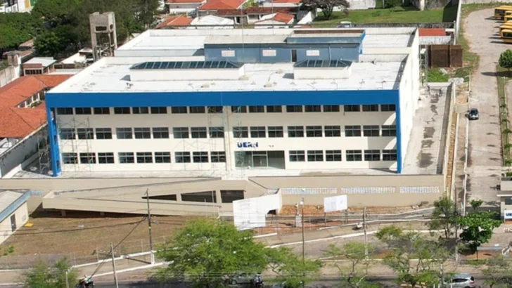 Vista aérea do campus da Uern Natal, com prédio de fachada branca e detalhes azuis, cercado por ruas, árvores e imóveis residenciais
