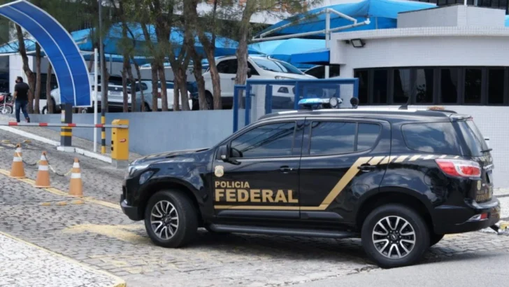 Viatura preta da Polícia Federal estacionada em frente a uma unidade com fachada branca, portão azul e veículos estacionados na área interna