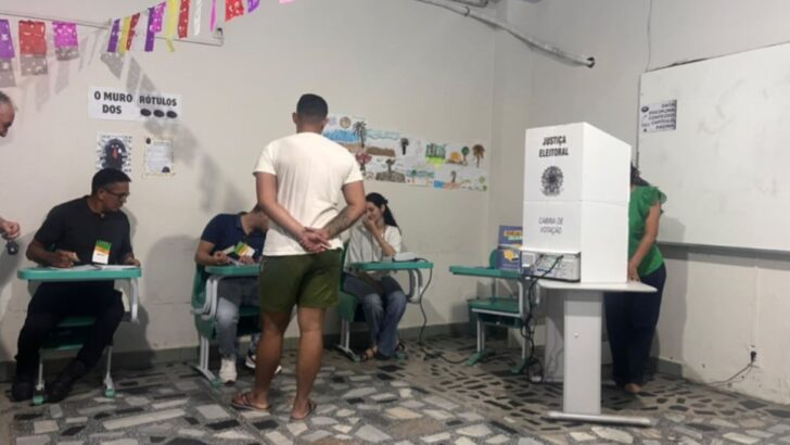 Eleitor aguarda diante de cabine de votação enquanto mesários trabalham em seção eleitoral