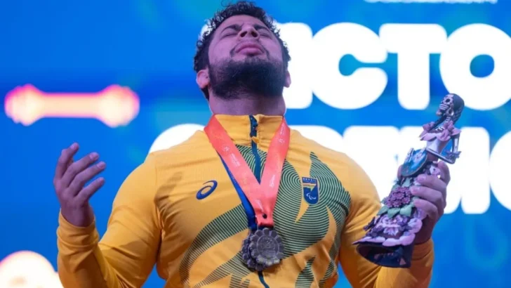 Atleta de halterofilismo com uniforme do Brasil, medalha no peito e troféu nas mãos durante cerimônia de premiação