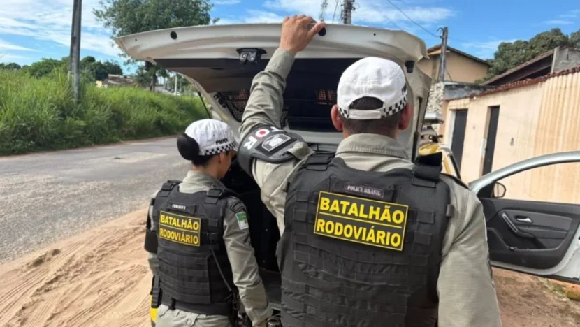 Dois policiais militares do Batalhão Rodoviário, vistos de costas e usando coletes de proteção, estão junto ao compartimento traseiro aberto de uma viatura estacionada em uma rua