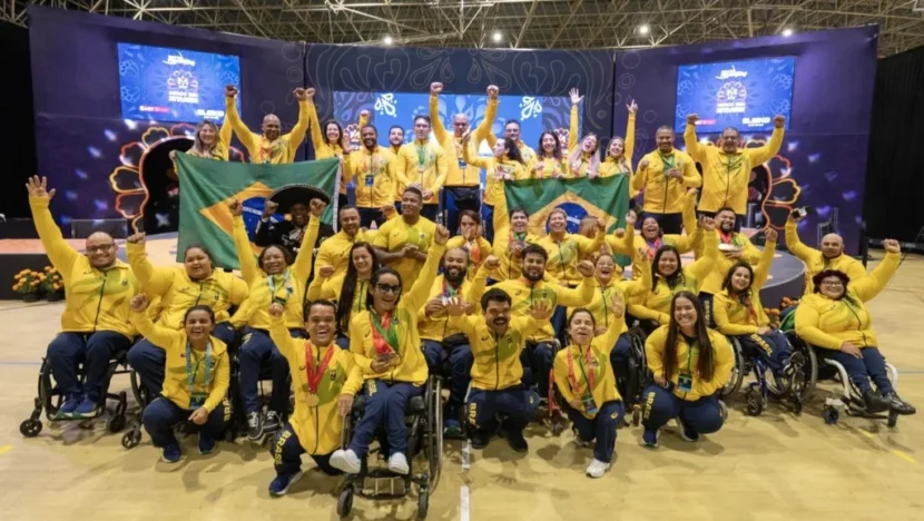 Grupo de atletas e integrantes da delegação brasileira de halterofilismo paralímpico posa em ginásio, usando uniformes amarelos e azuis, exibindo medalhas e bandeiras do Brasil. Parte dos atletas utiliza cadeiras de rodas