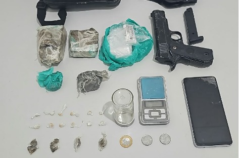 Materiais dispostos sobre uma superfície clara, incluindo embalagens com substâncias apreendidas, pequenas porções embaladas, objeto semelhante a uma pistola, carregador, balança digital, celular e moedas