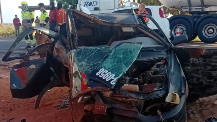 Automóvel escuro com a carroceria retorcida, teto deformado, para-brisa quebrado e parte dianteira destruída. Ao fundo, profissionais de atendimento e outras pessoas estão próximos a veículos de grande porte