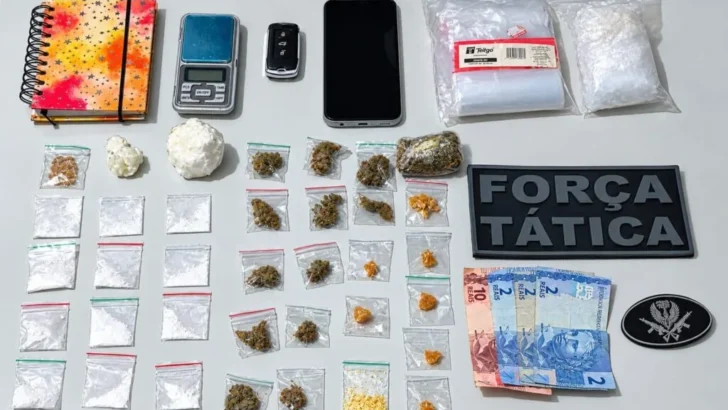 Materiais dispostos sobre uma mesa, incluindo pequenas embalagens com substâncias de diferentes cores, balança digital, telefone celular, caderno, notas de dinheiro e identificação da Força Tática