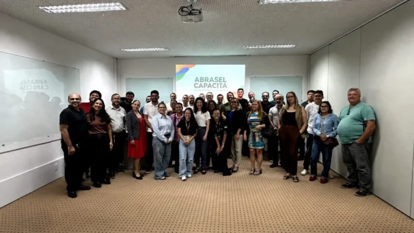 Grupo de participantes posa para fotografia em uma sala de eventos, diante de uma projeção com a inscrição “Abrasel Capacita”