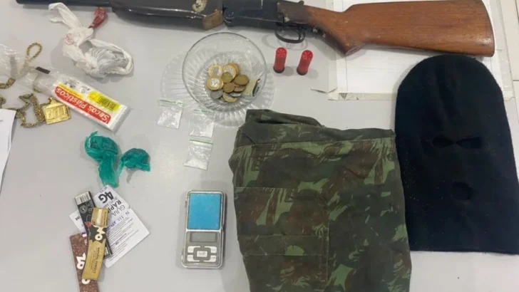 Espingarda de cano longo com coronha de madeira, dois cartuchos vermelhos, moedas, pequenas embalagens, balança digital, roupa camuflada e balaclava preta estão dispostos sobre uma mesa branca