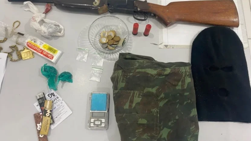 Espingarda de cano longo com coronha de madeira, dois cartuchos vermelhos, moedas, pequenas embalagens, balança digital, roupa camuflada e balaclava preta estão dispostos sobre uma mesa branca