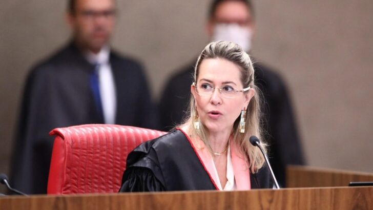 Advogada Maria Claudia Bucchianeri, de óculos e vestes jurídicas pretas, fala diante de um microfone, sentada em cadeira vermelha, durante sessão em ambiente judicial