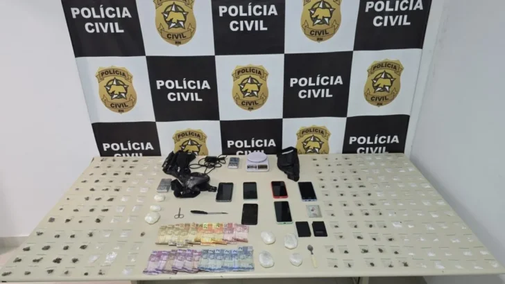 Mesa com diversas pequenas embalagens contendo materiais apreendidos, cédulas de dinheiro, celulares, balanças digitais e outros objetos, diante de painel com símbolos da Polícia Civil do Rio Grande do Norte