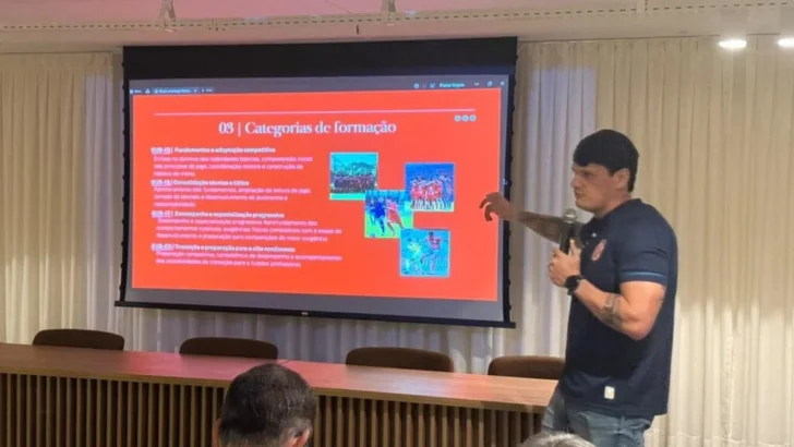 Homem com camisa azul-escura segura um microfone e aponta para uma apresentação projetada em tela vermelha, durante reunião em auditório com mesa e cadeiras
