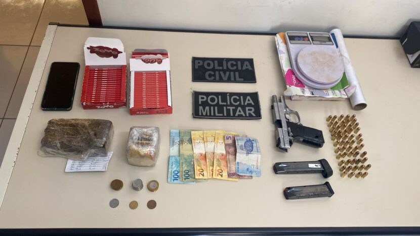 Mesa com pistola, dois carregadores, dezenas de munições, pacotes de material apreendido, cédulas e moedas, celular, balança digital e embalagens, ao lado de identificações da Polícia Civil e da Polícia Militar