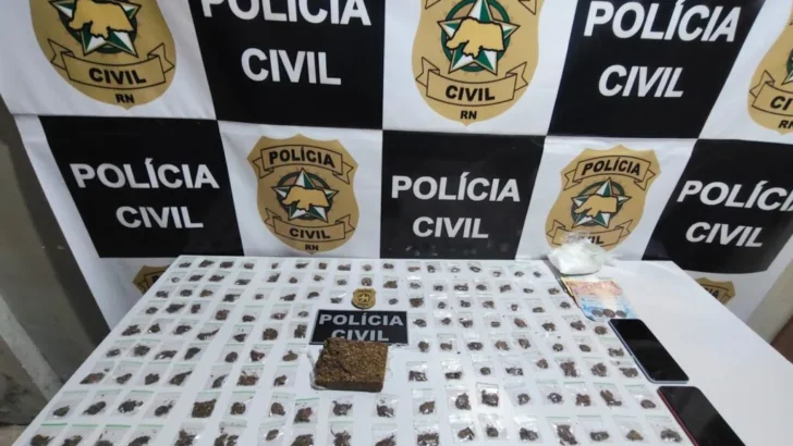 Mesa com dezenas de pequenas embalagens contendo material vegetal, uma porção maior da mesma substância, cédulas de dinheiro e dois celulares, diante de painel com símbolos da Polícia Civil do Rio Grande do Norte