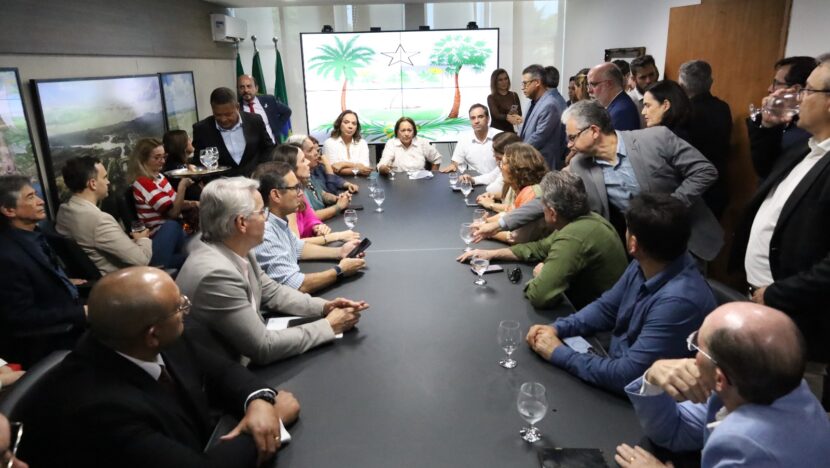 Fátima Bezerra e Cadu Xavier sentados à cabeceira de uma mesa cercada por participantes da reunião, com o brasão do Rio Grande do Norte ao fundo