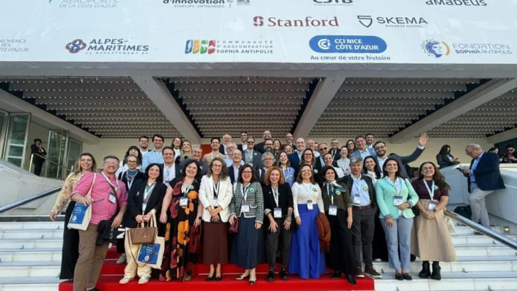 Grupo de participantes de uma conferência internacional posa para fotografia em uma escadaria com tapete vermelho. Ao fundo, aparecem logotipos de instituições e empresas ligadas à inovação, como Stanford, SKEMA e Sophia Antipolis