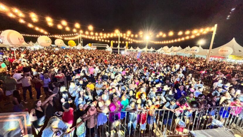 Multidão acompanha evento noturno em espaço aberto, iluminado por cordões de luzes, com tendas, estruturas de apoio e grades de proteção próximas ao palco
