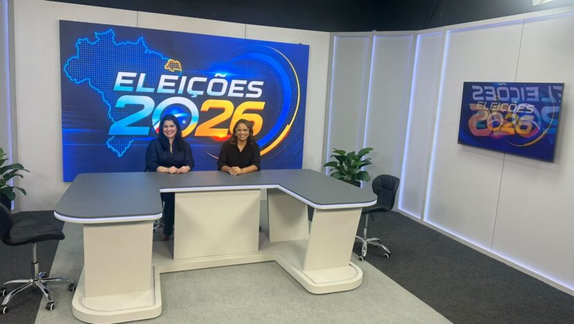 Duas mulheres atrás da bancada de um estúdio de televisão com painéis das Eleições 2026 ao fundo