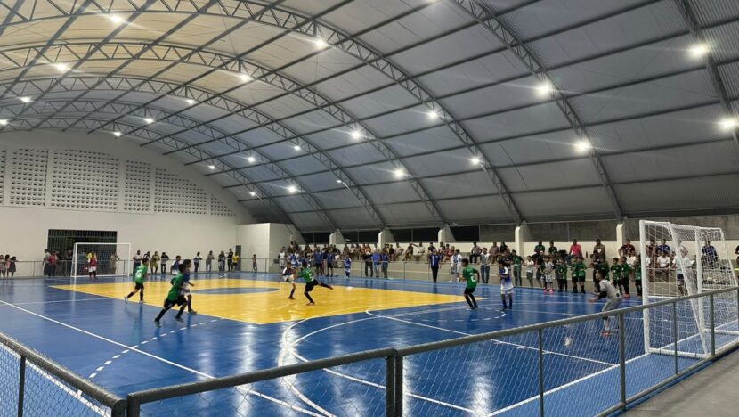 Jogadores em quadra azul e amarela de ginásio coberto, com público junto ao alambrado