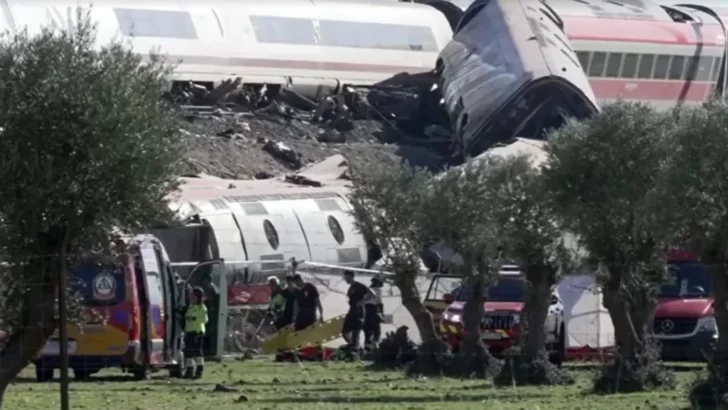 Vagões de trem descarrilados e danificados aparecem espalhados por uma área próxima aos trilhos, enquanto equipes de resgate trabalham no local com ambulâncias e veículos de emergência