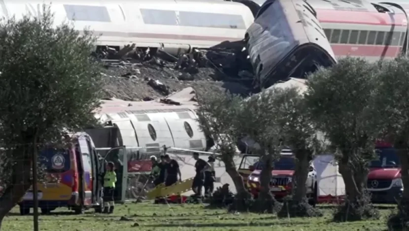 Vagões de trem descarrilados e danificados aparecem espalhados por uma área próxima aos trilhos, enquanto equipes de resgate trabalham no local com ambulâncias e veículos de emergência