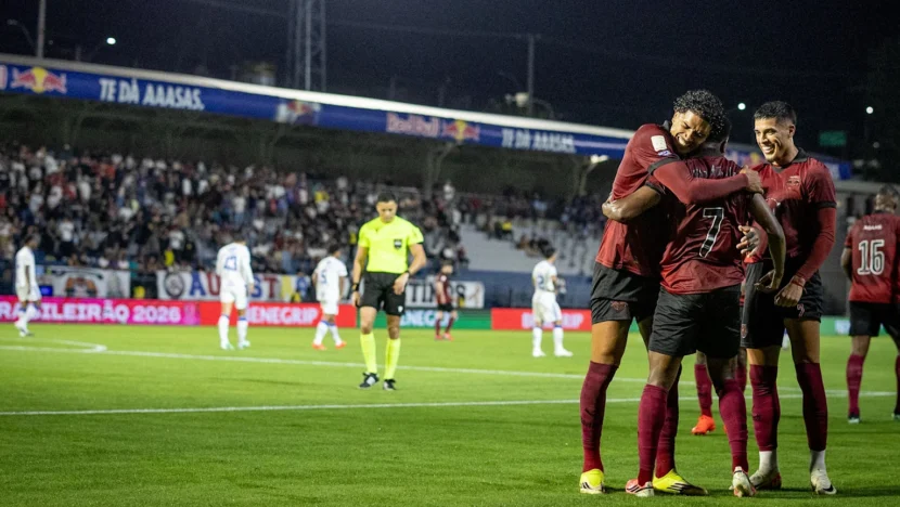 Eric Ramires, Lucas Barbosa e Guzmán Rodríguez se abraçam no gramado com uniformes vermelhos do Bragantino