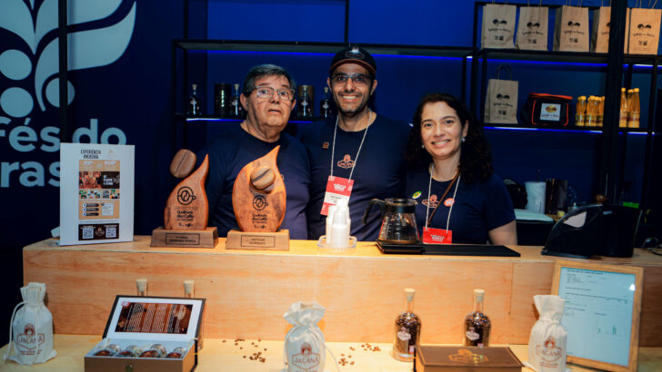 Três pessoas no estande do Café Jaçanã, com dois troféus sobre o balcão e produtos expostos