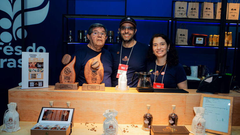 Três pessoas no estande do Café Jaçanã, com dois troféus sobre o balcão e produtos expostos