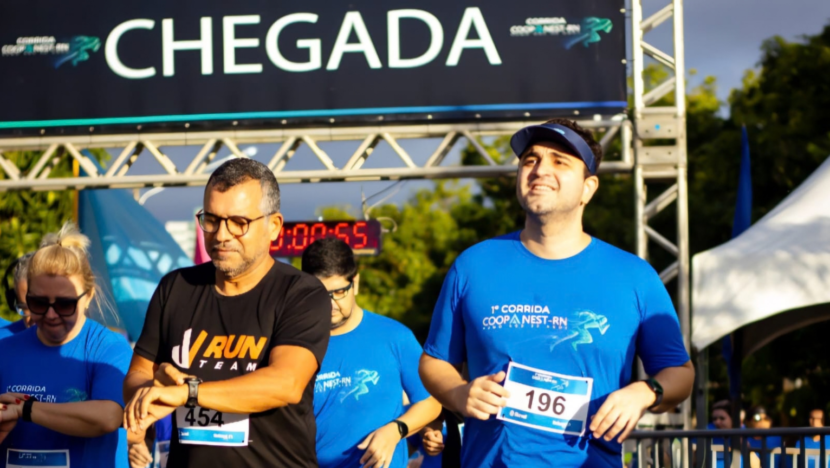 Corredores com camisetas azuis e números de peito passam por estrutura de chegada durante evento esportivo.