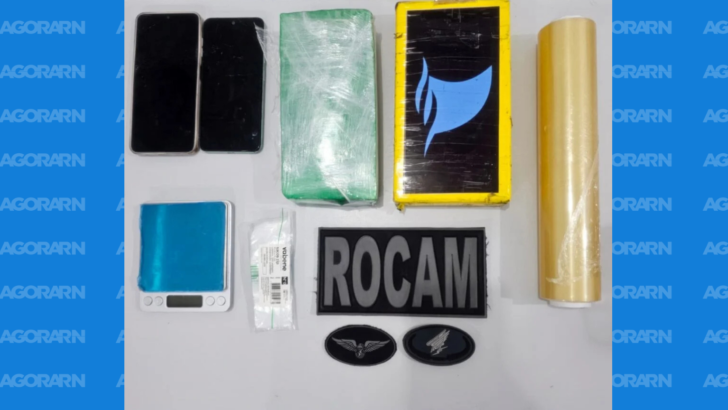 Materiais dispostos sobre uma superfície branca, incluindo dois celulares, dois pacotes retangulares embalados, balança digital azul, embalagens plásticas, rolo de plástico-filme e identificação da Rocam