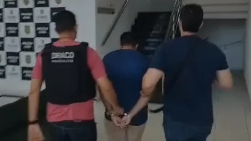 Imagem mostra um homem de camisa azul e bermuda clara, com as mãos algemadas para trás, sendo conduzido por dois homens em direção a uma escadaria no interior de um prédio
