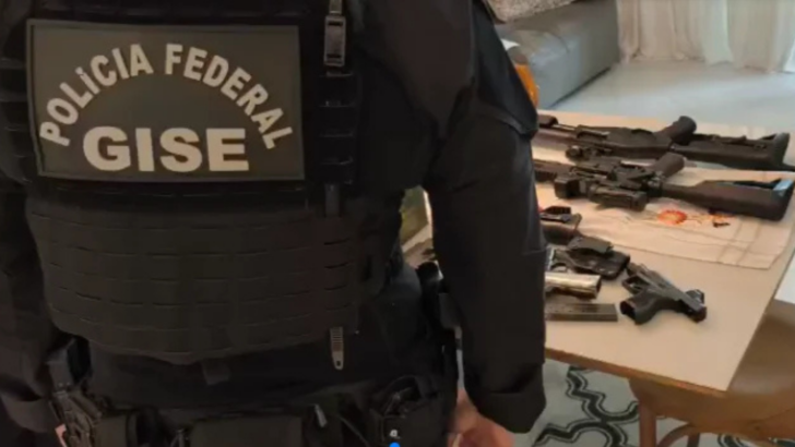Agente com uniforme e identificação da Polícia Federal e do GISE aparece de costas diante de uma mesa com armas de fogo durante ação policial