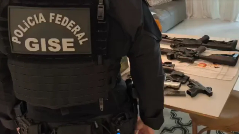 Agente com uniforme e identificação da Polícia Federal e do GISE aparece de costas diante de uma mesa com armas de fogo durante ação policial